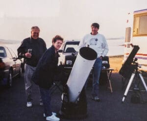 Three people standing around a telescope on the ground.